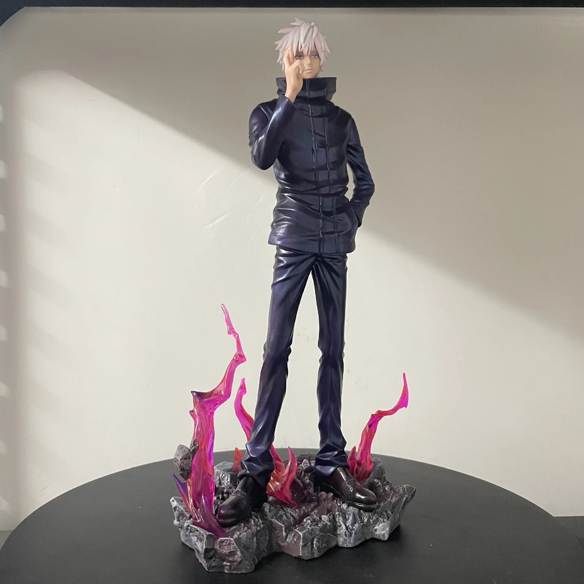Figurine en tenue noire, cheveux blancs, pose près d’un socle avec flammes roses.