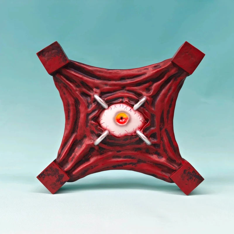 Figurine Œil Maudit rouge avec œil orange, orbite blanche et quatre poignées carrées.